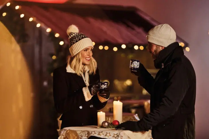 Ein Mann und eine Frau stehen in einer winterlichen Umgebung mit einer kleinen Bude und Lichterketten an einem Stehtisch, unterhalten sich und trinken Punsch. 