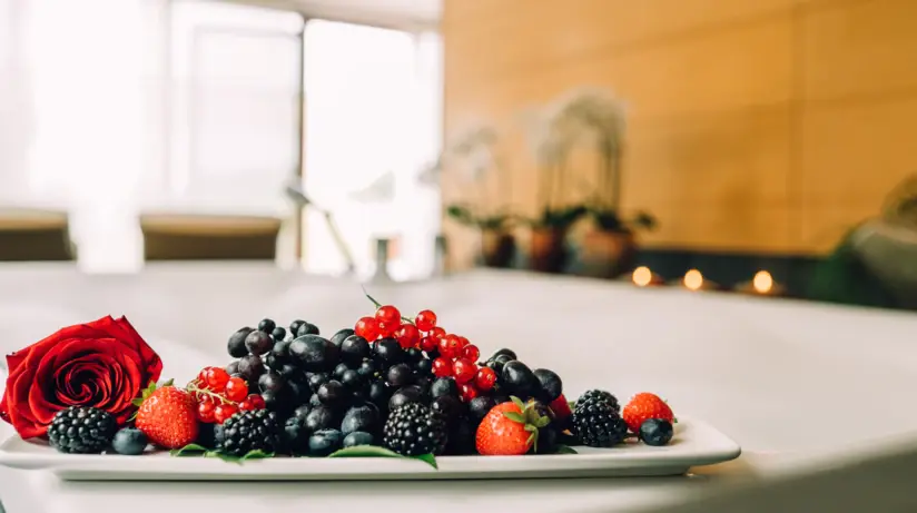 Ein Teller, angerichtet mit Erdbeeren, Blaubeeren, Brombeeren und Johannisbeeren sowie einer Rose, ist auf einem Badewannenrand abgestellt. Im Hintergrund ist verschwommen ein SPA-Bereich zu erkennen. 