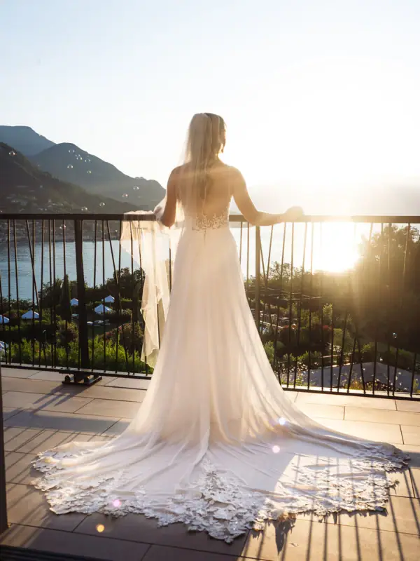 Eine Braut im weißen Kleid steht auf einer Terrasse und blickt auf den Gardasee im Abendlicht.