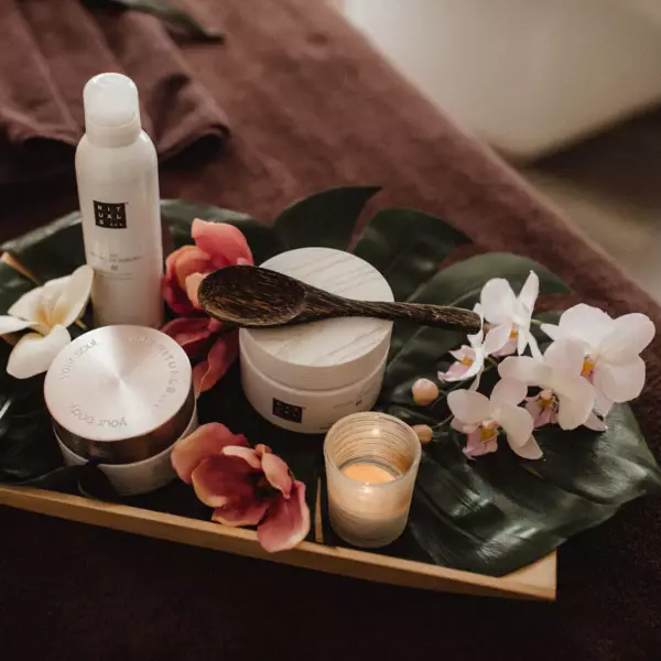  Ein Tablett mit Spa-Produkten, darunter eine Lotionflasche, ein Tiegel mit Creme, eine Kerze, eine Holzkelle und frische Orchideenblüten, arrangiert auf einem großen grünen Blatt und platziert auf einem braunen Handtuch.