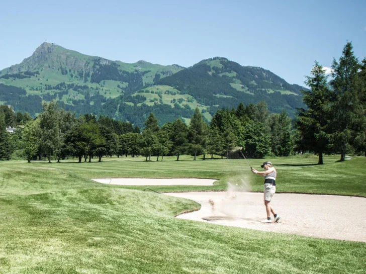 Eine Frau schlägt einen Golfball auf einem sandigen teil des Golfplatzes und wirbelt en Sand durch die Luft. Im Hintergrund liegen die grünen Berge und die Sonne scheint warm. 