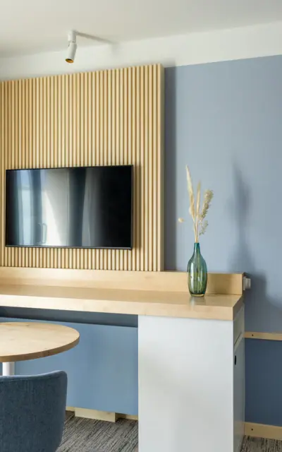 Modernes Zimmerinterieur mit einer Wand aus Holzlamellen, an der ein Flachbildfernseher montiert ist. Unter dem Fernseher befindet sich eine schlichte, helle Holzkonsole mit einer stilvollen, blau-grünen Glasvase, die Trockenblumen enthält. Die Wand dahinter ist in einem sanften Blauton gestrichen, was eine ruhige und entspannende Atmosphäre schafft.