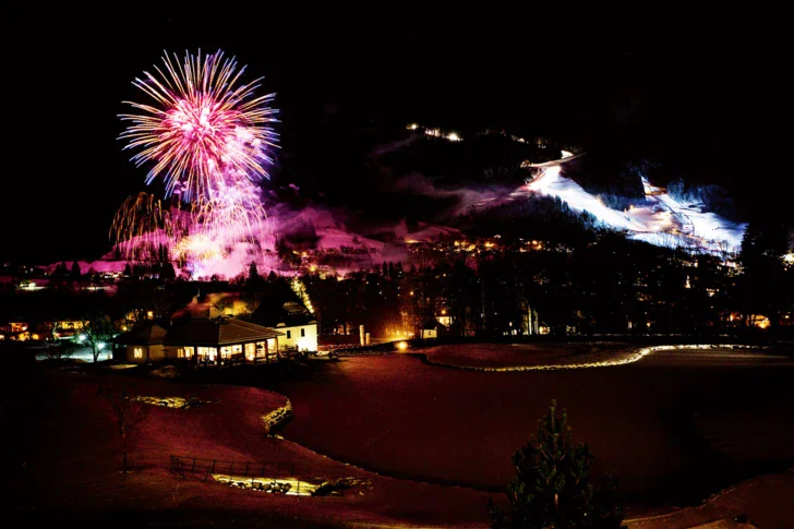 Farbenprächtiges Feuerwerk explodiert über einer nächtlichen Winterlandschaft mit beleuchteten Skipisten im Hintergrund und einer Resortlandschaft im Vordergrund. 
