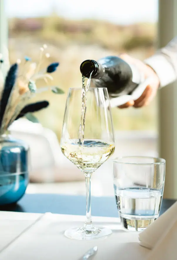 Ein Kellner gießt Weißwein in ein Weinglas an einem elegant gedeckten Tisch im Dünenrestaurant. Der Hintergrund ist verschwommen, bietet jedoch eine helle und natürliche Umgebung, vermutlich mit Aussicht auf Dünen oder Strand. Auf dem Tisch sind das Weinglas und ein Wasserglas zu sehen, daneben eine stilvolle Tischdekoration mit einem blauen Vasenarrangement. Der Fokus liegt auf der klaren Bewegung des Weines und der professionellen Servicegeste.