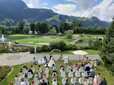 Eine Trauungszeremonie auf einer grünen Wiese im Hotel Kitzbühel. Im Hintergrund befindet sich eine Außenlandschaft aus Gewässern, Steinbrücken und Wiesen sowie Bergen. 
