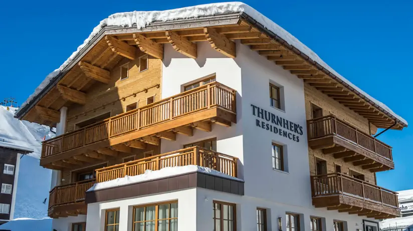 An alpine chalet with a mixture of white and wooden exterior walls and wooden balconies. The lettering of Thurnher's Residences can be seen on the outer wall.