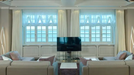 Ein helles Wohnzimmer in einer Suite mit einem Fensterblick auf die Ostsee. Die Sofas sind in neutralen Farbtönen gehalten und mit dekorativen Kissen in Rosa- und Violettnuancen versehen. Vor den klassischen weißen Fensterrahmen hängen elegante, weiße Vorhänge und transparente, helle Gardinen. Zwischen den Sofas steht ein moderner Flachbildfernseher auf einem Glasständer, umgeben von kreativen Stehlampen, die eine gemütliche Beleuchtung bieten.