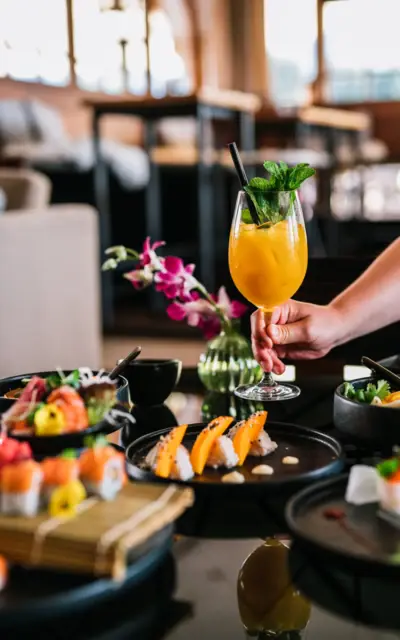 Ein erfrischender Cocktail in einem eleganten Glas wird in einem stilvollen Sushi-Restaurant serviert. Der Fokus liegt auf dem orangefarbenen Getränk, das mit Minze garniert ist, und im Hintergrund sind kunstvoll arrangierte Sushi-Platten und eine asiatisch inspirierte Tischdekoration zu sehen, was eine exklusive und genussvolle Atmosphäre vermittelt.