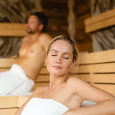 Eine Frau sitzt in einer Sauna, ein Mann im Hintergrund.