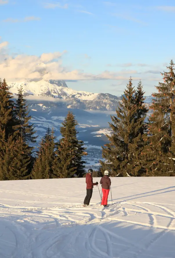 Zwei Skifahrer stehen inmitten einer schneebedeckten Landschaft, umgeben von hohen Tannen. Sie blicken auf eine atemberaubende Bergkulisse mit einem majestätischen, teilweise wolkigen Gipfel in der Ferne. Der Himmel ist blau und mit sanften Wolken in ein warmes Licht getaucht. 