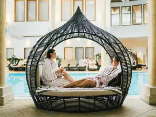 Zwei Frauen in weißen Bademänteln entspannen in einer stilvollen Rattan-Lounge am Rand eines luxuriösen Indoor-Pools, genießen Getränke und eine ruhige Wellness-Atmosphäre.