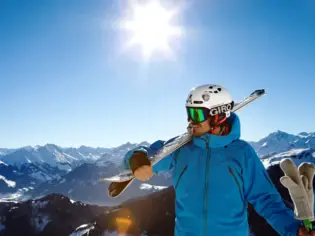 Ein Mann in blauer Jacke steht auf einem Berg und hat seine Skier auf der Schulter. Im Hintergrund erstreckt sich eine Bergkulisse und die Sonne scheint. 