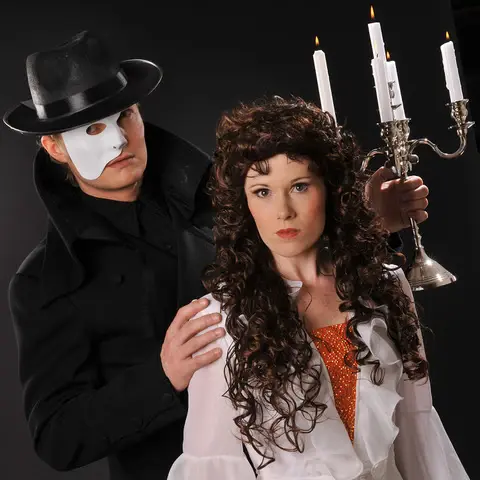 Ein Mann in schwarzem Mantel und Hut mit weißer Maske, neben einer Frau mit braunen Locken und einem Kronleuchter in der Hand – inspiriert vom "Phantom der Oper".