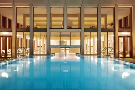 Einladender Poolbereich bei warmem Licht in einem Luxushotel. Die ruhige Wasseroberfläche des blauen Pools spiegelt die umgebende, moderne Architektur und Beleuchtung wider. Liegestühle und Entspannungsbereiche sind symmetrisch angeordnet und bieten einen Blick auf die ruhige und entspannte Atmosphäre des SPA-Bereichs bei Nacht.