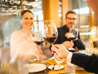 Eine Gruppe von Menschen stößt mit Rotweingläsern in einer eleganten Restaurantatmosphäre an, während eine Frau und ein Mann im Hintergrund lächelnd miteinander anstoßen.