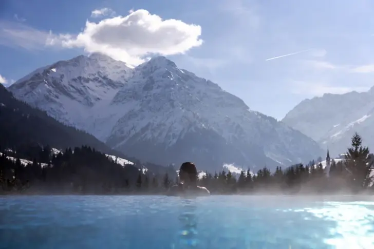 Eine Frau schwimmt in einem warm Dampfenden Pool und überblickt die schneebedeckten Berge bei Sonnenschein. 