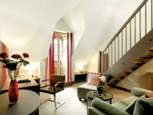 Ein Wohnbereich mit zwei Sesseln, einem kleinen Glastisch, einem Schreibtisch und einem Stuhl. Vorne links im Bild ist eine rote Vase mit Rosen zu sehen und im Hintergrund führt eine Holztreppe nach oben.