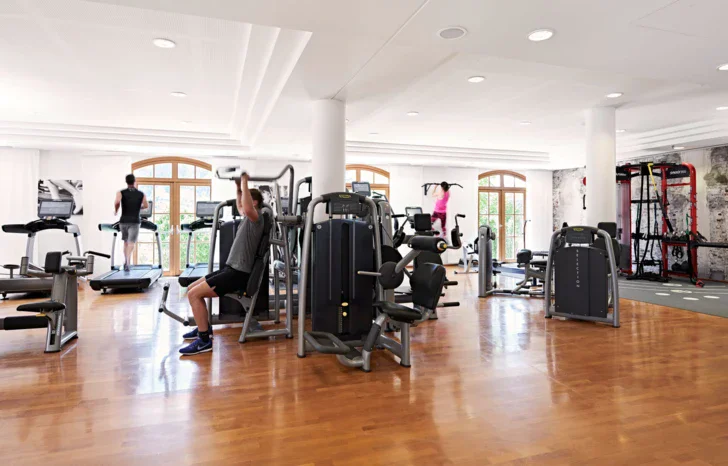 Fitnessraum mit Holzboden und weißen Wänden worin unterschiedliche Geräte stehen, mit denen trainiert wird.  