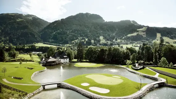 Ein Golfplatz mit einem See und einem Haus im Hintergrund, umgeben von Bäumen und Bergen.