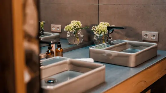 Ein Waschtisch mit zwei Waschbecken in grau. In der hinteren Ecke steht eine kleine Vase mit Blumen und es sind zwei Seifenspender zu sehen. 
