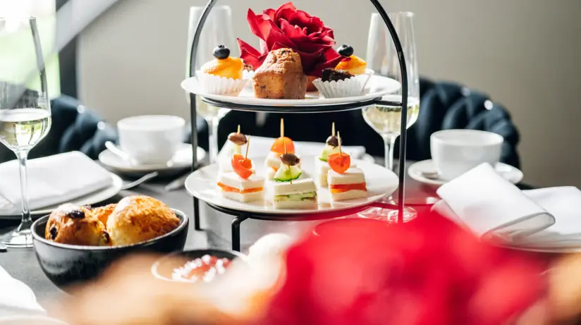 Ein Esstisch mit einem Etagere an kleinen Häppchen und Gebäck, neben dem Kaffeetassen, Sektgläser und Teller verteilt sind. 