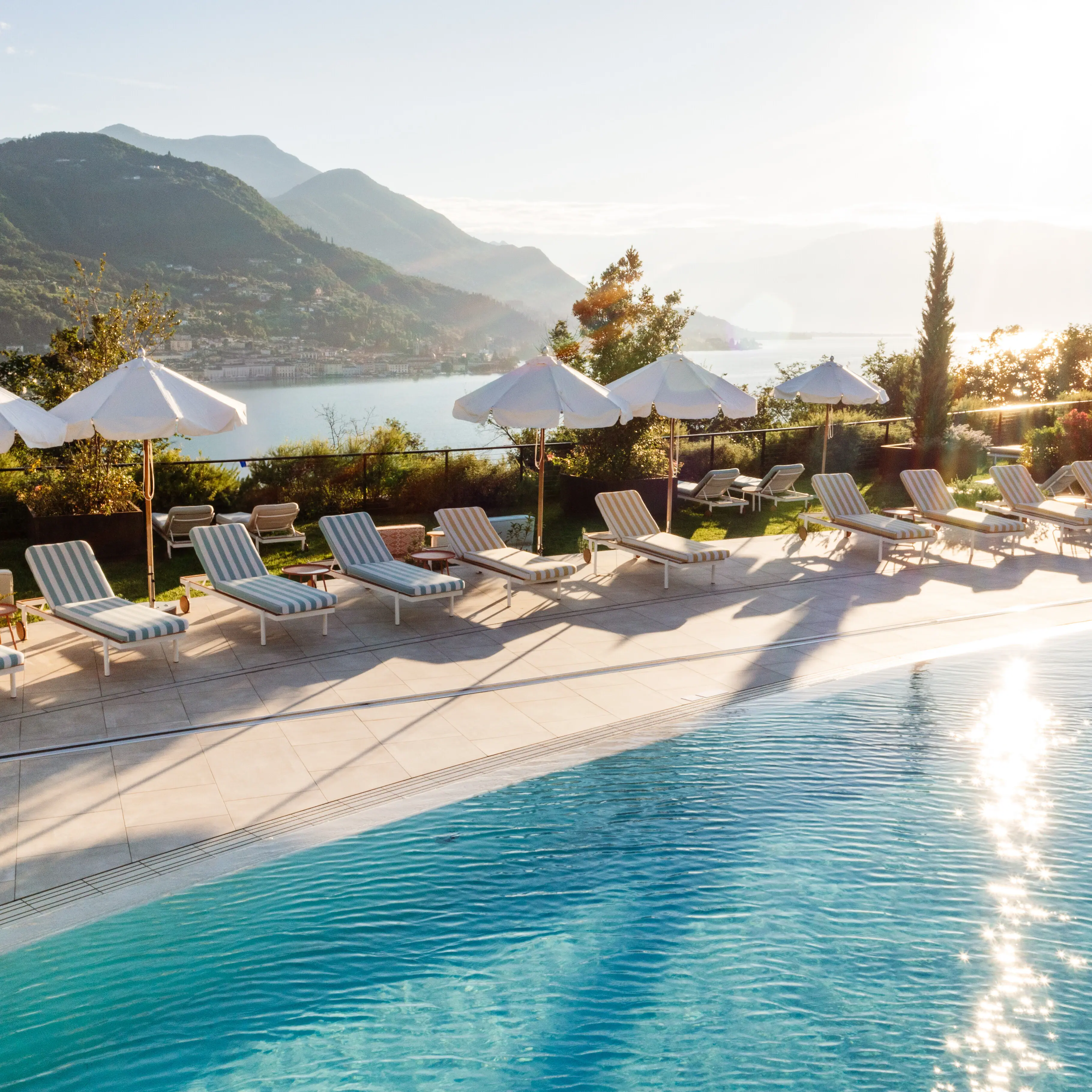 A-ROSA Gardasee Ein Pool mit Liegestühlen und Sonnenschirmen an einem Gewässer.