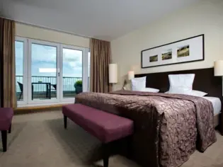 Ein großes Bett mit lilaner tagesdecke steht in einem geräumigen Hotelzimmer. An der Wand hängt ein großes Bild über dem Kopfteil und im Hintergrund ist eine große Fensterfront mit Zugang zu einer Terrasse zu sehen.