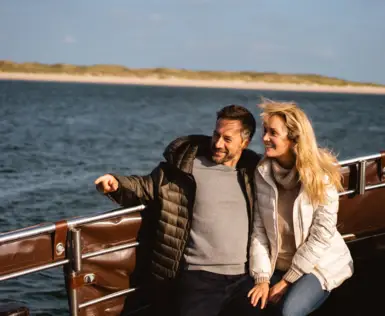Ein Paar sitzt Arm in Arm, lächelnd auf einem offenen Boot. Der Mann hat seinen Finger auf etwas außerhalb des Bildes gerichtet. im Hintergrund sind Dünen und das Meer zu sehen. 