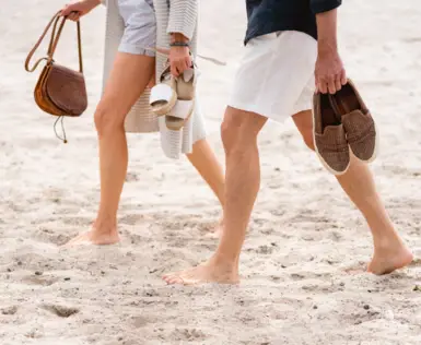 Ein Mann und eine Frau gehen barfuß im Sommeroutfit am Strand.