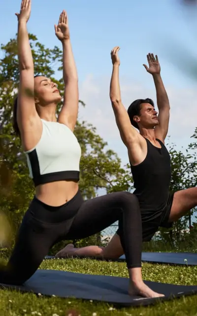 A man and a woman do the warrior pose on a yoga mat on a green meadow outdoors. 