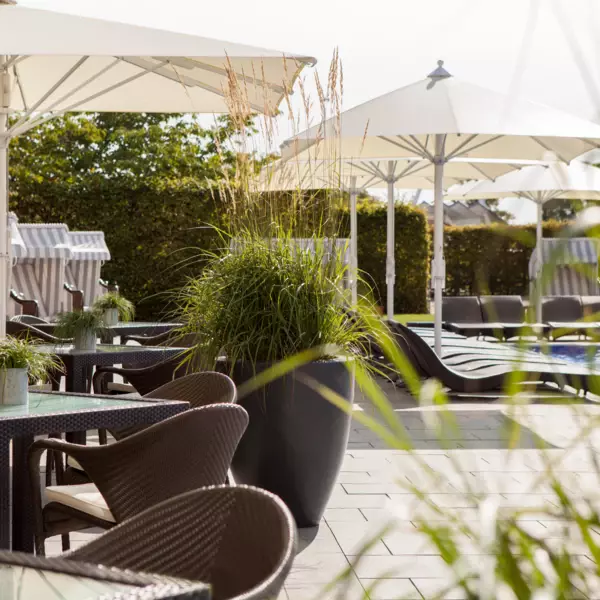 Ein Außenbereich eines Restaurants oder Cafés an einem sonnigen Tag mit Sonnenschirmen, gewebten Rattanmöbeln und großen Pflanzenkübeln, der an einen Poolbereich angrenzt.