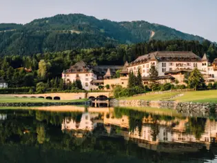  Ein traditionelles Resort, in weiß mit vielen Fenstern und dunklem Dach, spiegelt sich in einem ruhigen See vor der Kulisse eines dicht bewaldeten Berges in der Morgendämmerung.