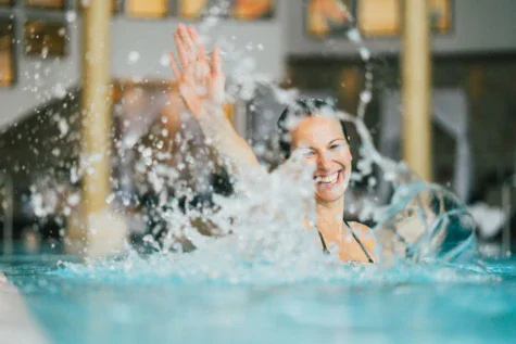 Eine Frau lächelt und klatscht ins Wasser, wodurch Spritzer in einem hell erleuchteten Innenpool entstehen. Im Hintergrund ist eine verschwommene Ansicht der Poolumgebung zu erkennen.