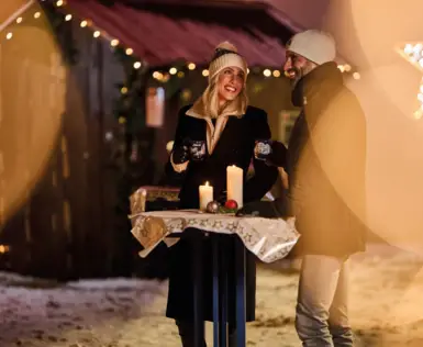 A man and woman standing outside holding candles, surrounded by festive lights, dressed in winter clothing.