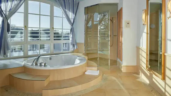 A luxurious bathroom with a spacious, all-round glazed corner bath. Bright daylight floods in through the white transom windows. Opposite the bath, a modern, see-through shower cubicle adds an elegant touch, while the golden fittings and handles add a touch of opulence. The warm tone of the tiles lining the bathroom creates a calming atmosphere, which is rounded off by the simple white towels and the soft light of a wall lamp.