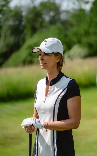 Frau mit weißer Kappe und weißem Hemd steht auf einer Wiese beim Golfspielen.