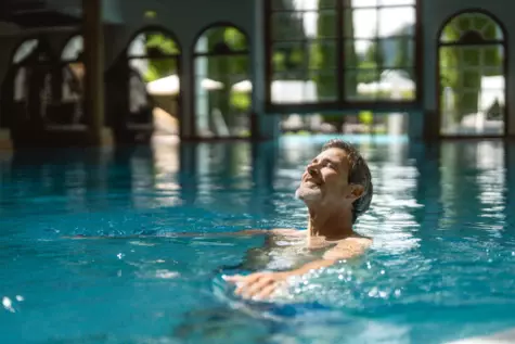Ein Mann genießt entspannt das Schwimmen in einem Innenpool, mit dem Gesicht nach oben gerichtet und den Augen geschlossen, im Hintergrund sind große Bogenfenster und Tageslicht zu sehen