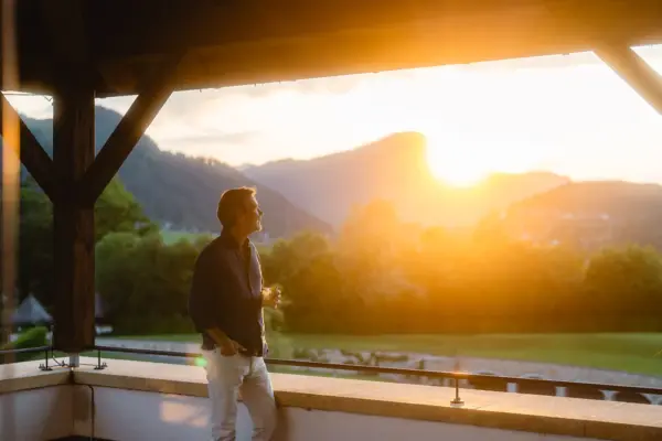 Ein Mann mit einem Glas in der Hand steht lässig auf einer Terrasse, die eine grüne Wiese, Wälder und Berge überblickt, über denen die Sonne aufgeht und in warmem orange leuchtet. 