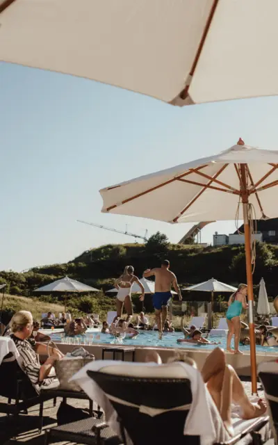 Gäste entspannen sich auf Sonnenliegen am Pool unter Sonnenschirmen, während andere Gäste im Hintergrund ins Wasser springen.