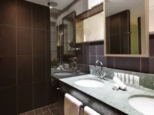 A bathroom with dark tiles, a shower and a vanity unit with two washbasins.