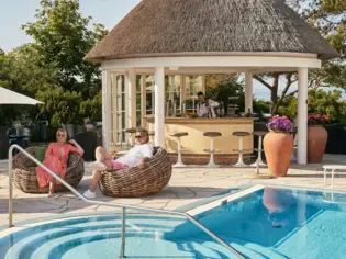 Ein Mann und eine Frau sitzen in Korbstühlen neben einem Swimmingpool und einer Poolbar.