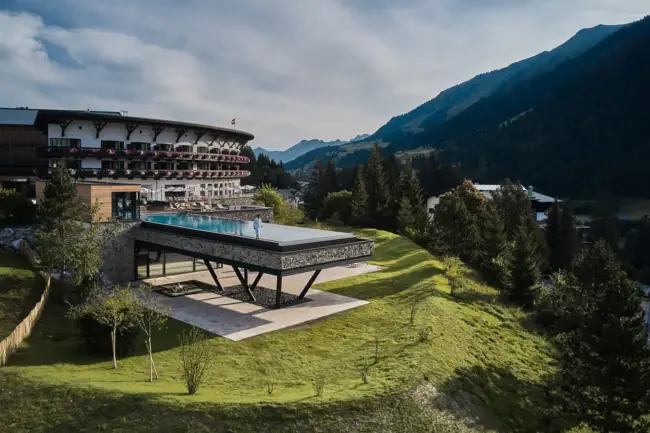 A-ROSA Ifen Hotel Kleinwalsertal Ein Gebäude mit einem Pool in der Mitte, umgeben von Bergen und blauem Himmel.