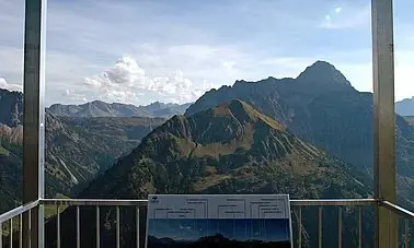 Infotafel auf einer Aussichtsplattform mit einer Berglandschaft mit leicht bewölktem Himmel im Hintergrund.