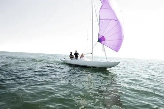 Drei Personen segeln bei sonnigem Wetter auf einem weißen Boot mit violettem Segel über ruhiges Meer.