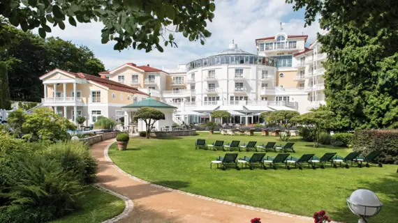Ein elegantes Hotel mit klassischer Architektur, umgeben von einer gepflegten Gartenanlage. Auf der grünen Wiese stehen mehrere Sonnenliegen.