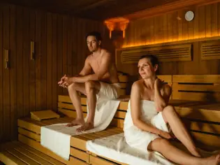 Ein Mann und eine Frau sitzen in einer Sauna auf Bänken und lächeln.