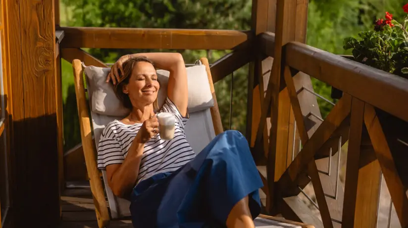 Eine Frau genießt in einem Liegestuhl die Sonne, welche auf den hölzernen Balkon fällt. 