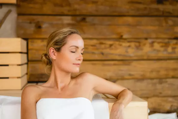 Eine Frau sitzt mit lockerem, blonden Dutt im Nacken in ein Handtuch gewickelt entspannt in der Sauna. Im Hintergrund ist die Holzverkleidung zu erkennen. 