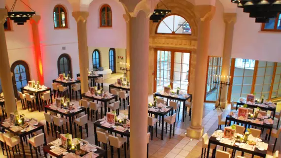 Das Bild zeigt einen elegant dekorierten Speisesaal mit mehreren Tischen, die für ein Abendessen vorbereitet sind. Die Tische sind mit weißen Tischdecken, Geschirr, Gläsern und kleinen Dekorationen ausgestattet, was auf ein bevorstehendes Event wie ein Bankett oder eine Gala hinweist. Die Beleuchtung ist warm und einladend, und die roten Akzente, wie der rote Teppich und die beleuchteten Säulen, geben dem Raum eine festliche Atmosphäre. Es ist ein repräsentativer Raum, der für feierliche Anlässe geeignet scheint.