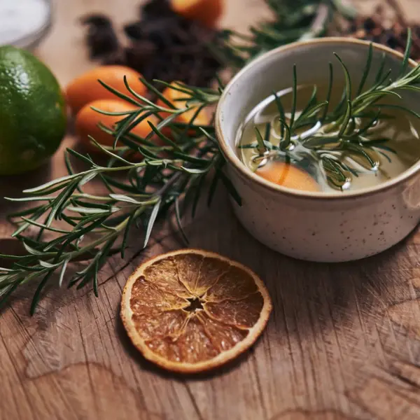 There is a bowl of oils on a wooden base and rosemary bushes, limes and oranges next to it. 